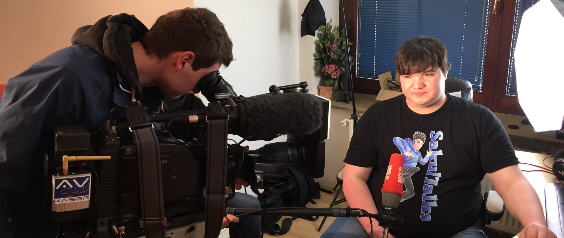 Lukas Krämer sitzt auf ein Stuhl vor ihm ist ein Kamerateam von SWR und Filmen ihn bei seiner Arbeit